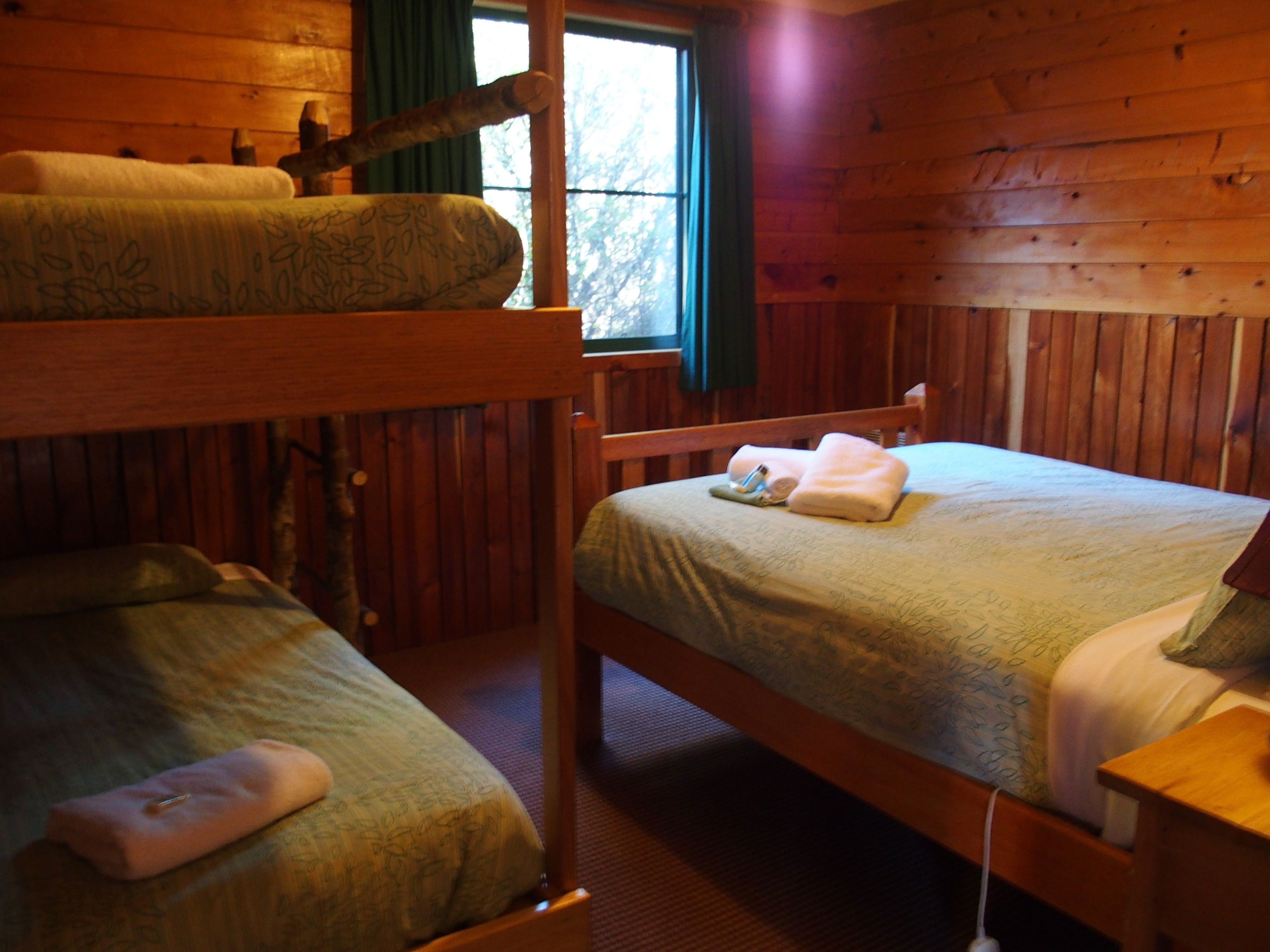 Una habitación tipo cabaña con dos literas, un cabecero de madera, una ventana con cortinas y una mesita de noche con lámpara.