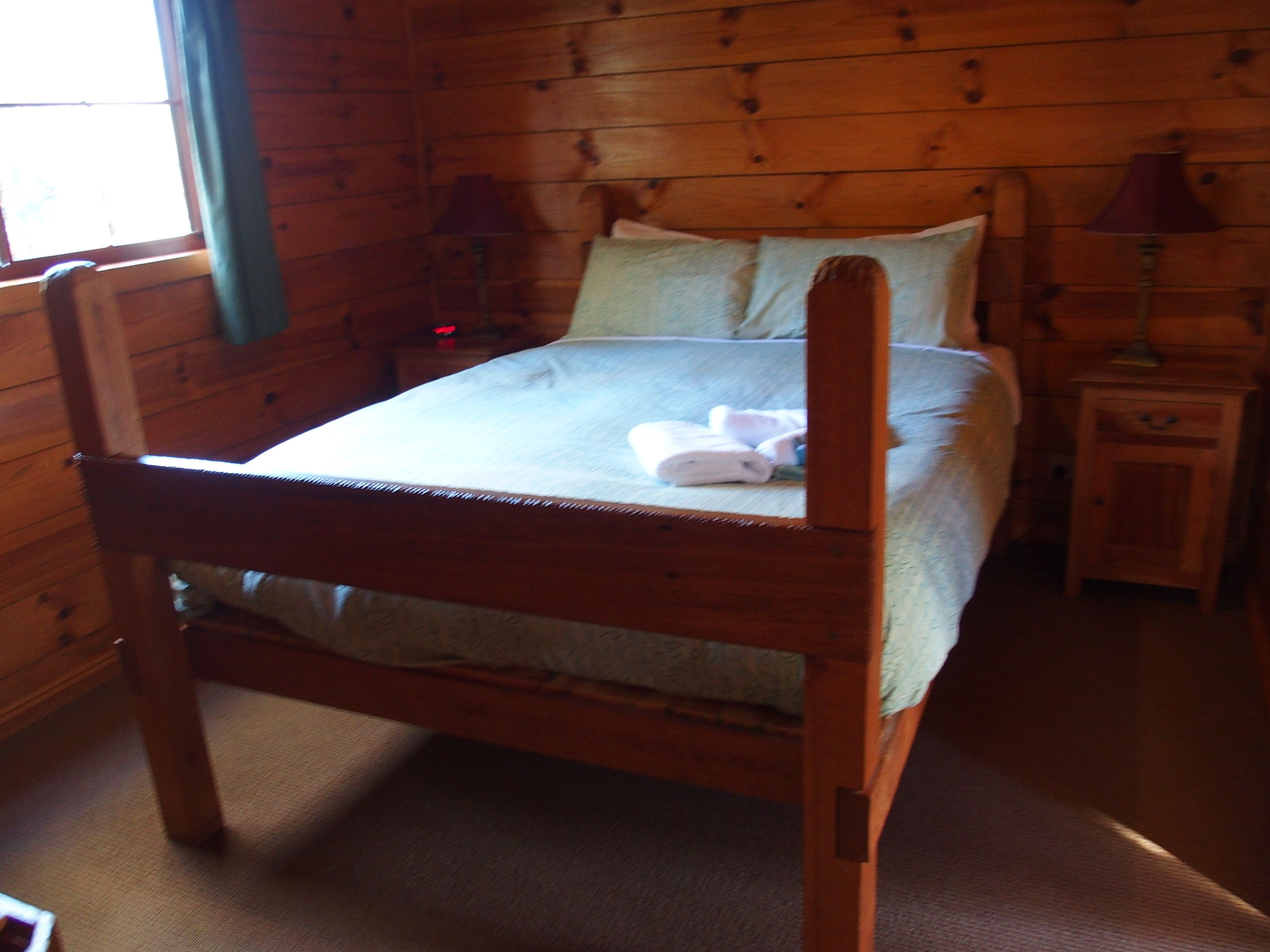 Una cama de madera en una habitación con ventana, mesita de noche y lámpara.