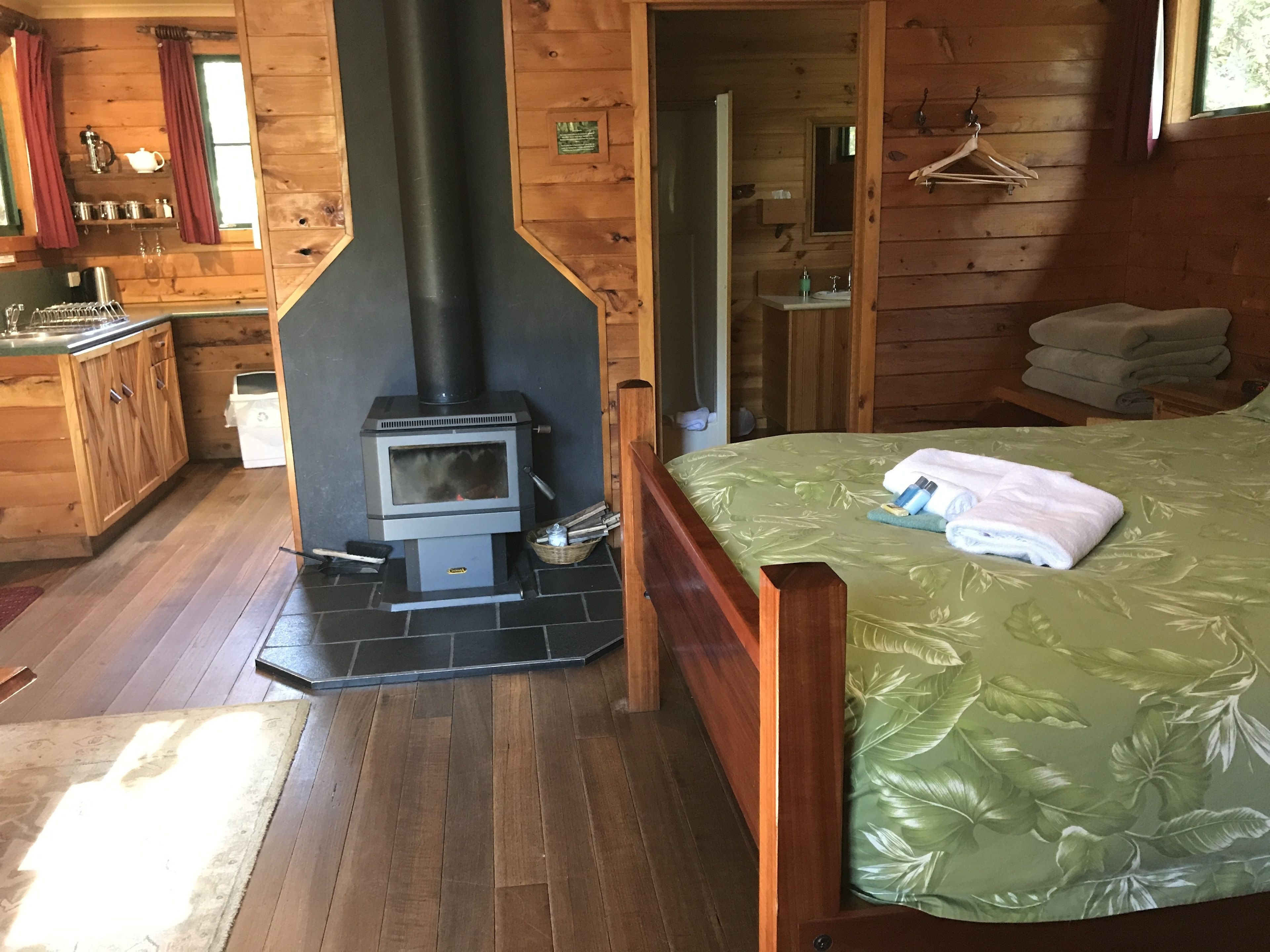 A wooden cabin interior with a central wood-burning stove, a bed with a green leaf-patterned bedspread, a wooden dresser, and a small bathroom with a mirror and sink.