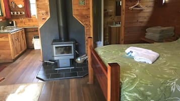 A wooden cabin interior with a central wood-burning stove, a bed with a green leaf-patterned bedspread, a wooden dresser, and a small bathroom with a mirror and sink.