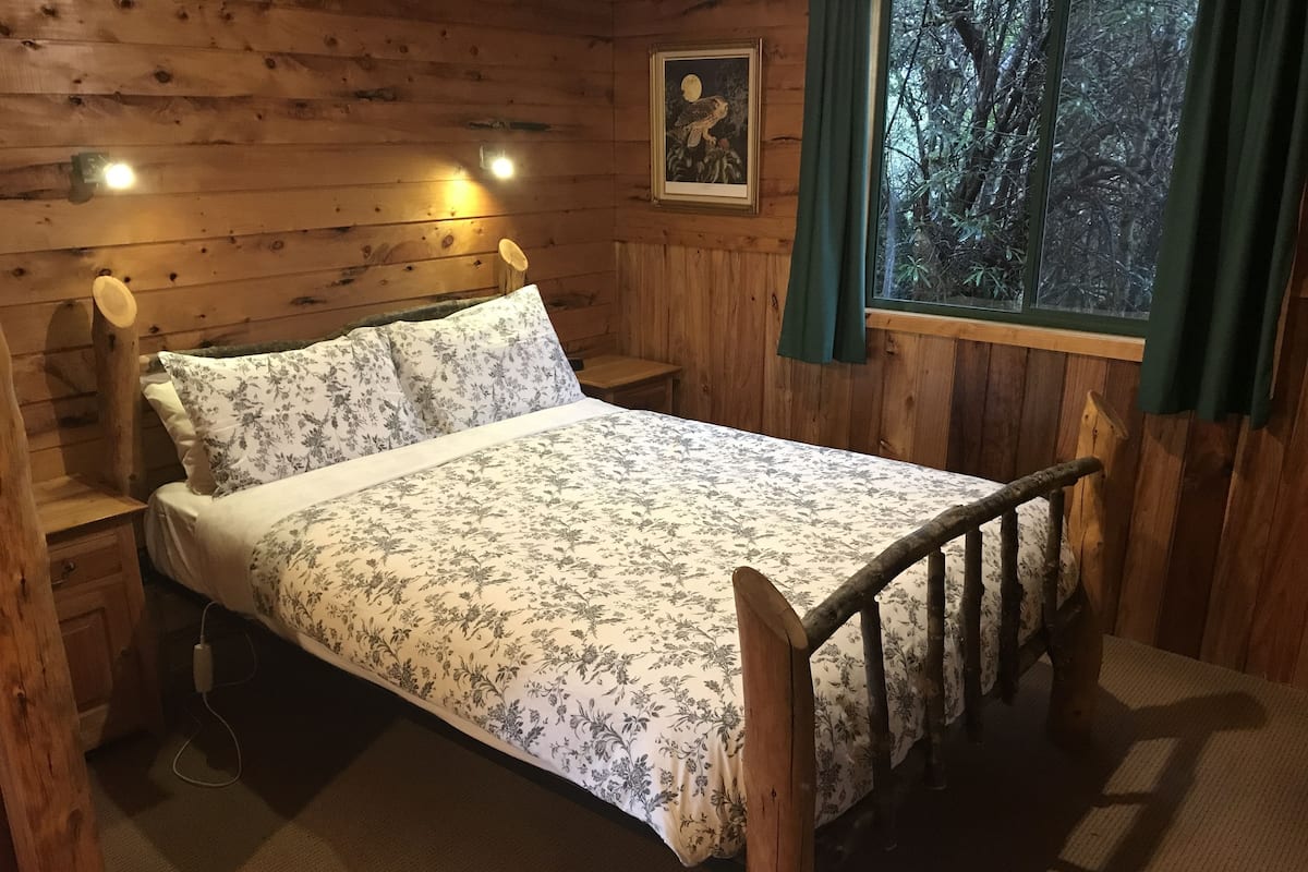 Un dormitorio de cabaña de madera con una cama, mesitas de noche y una ventana con vistas a árboles.