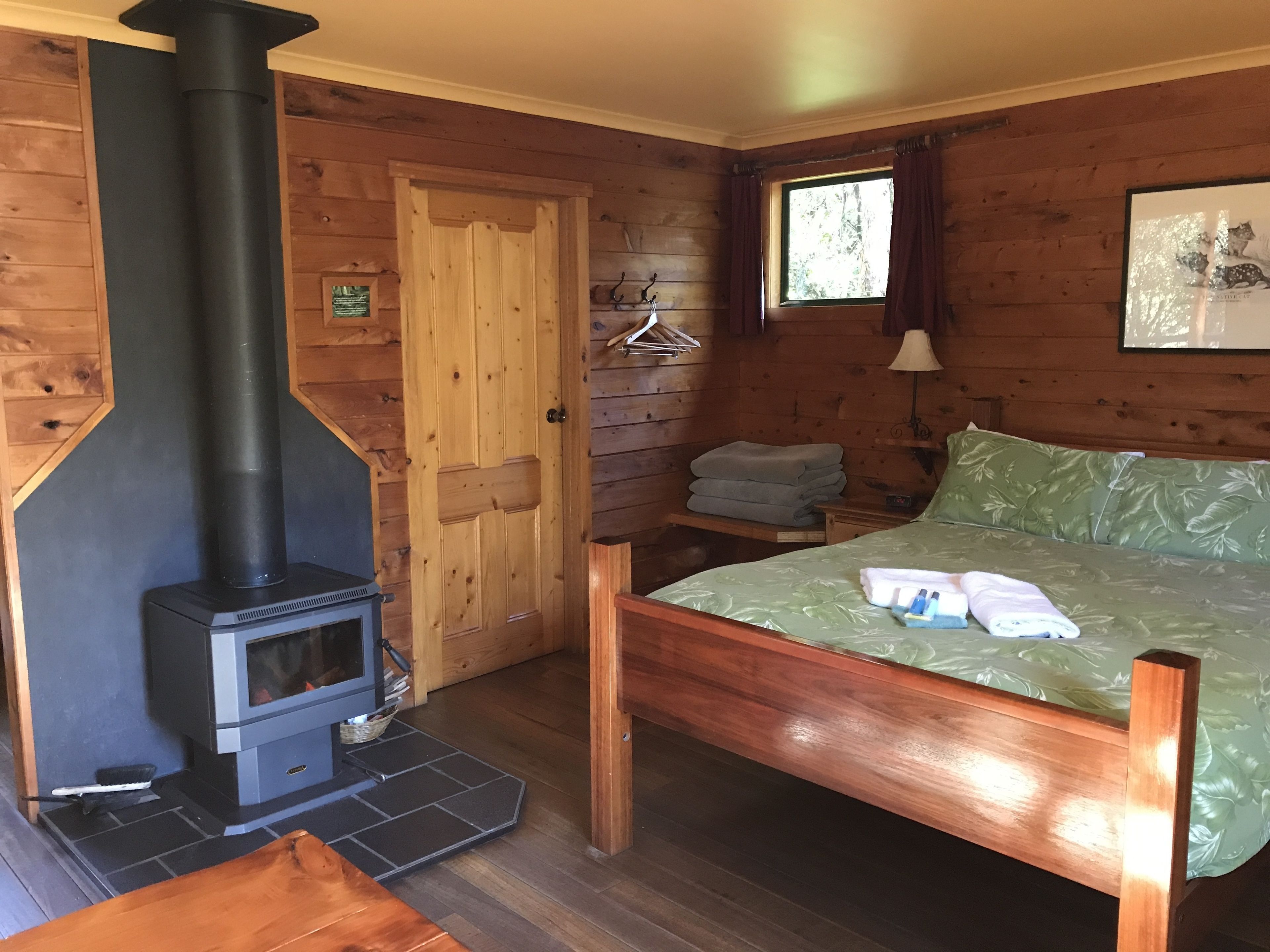 Habitación de cabaña de madera con una cama, un estufa de leña empotrada, una ventana con cortinas y una lámpara fijada a la pared.