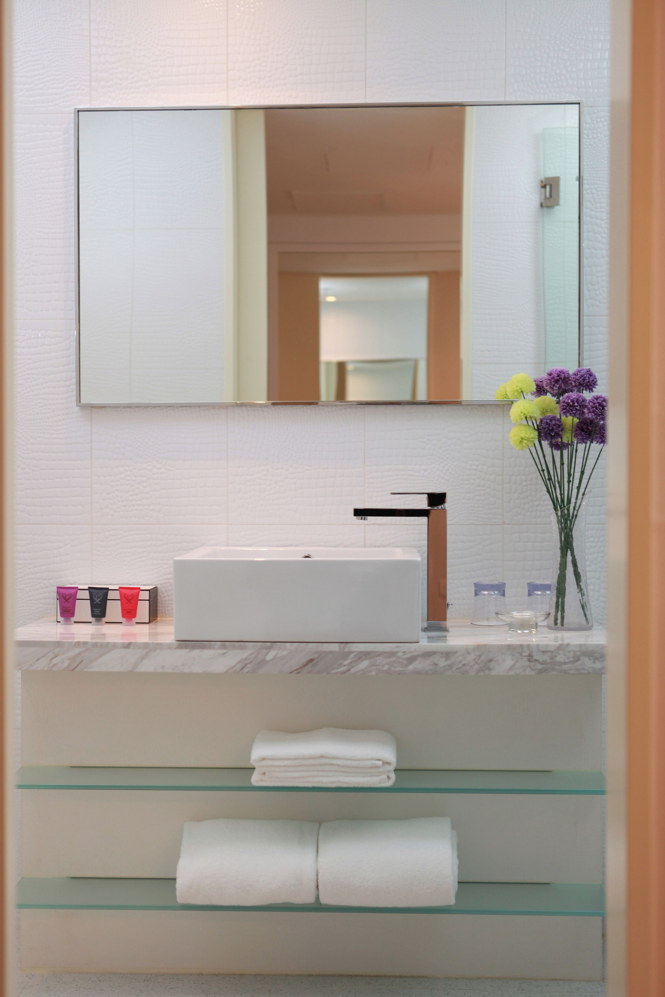 A bathroom with a white sink, a mirror, and a vase with flowers. There are towels on shelves and a glass partition below the sink.