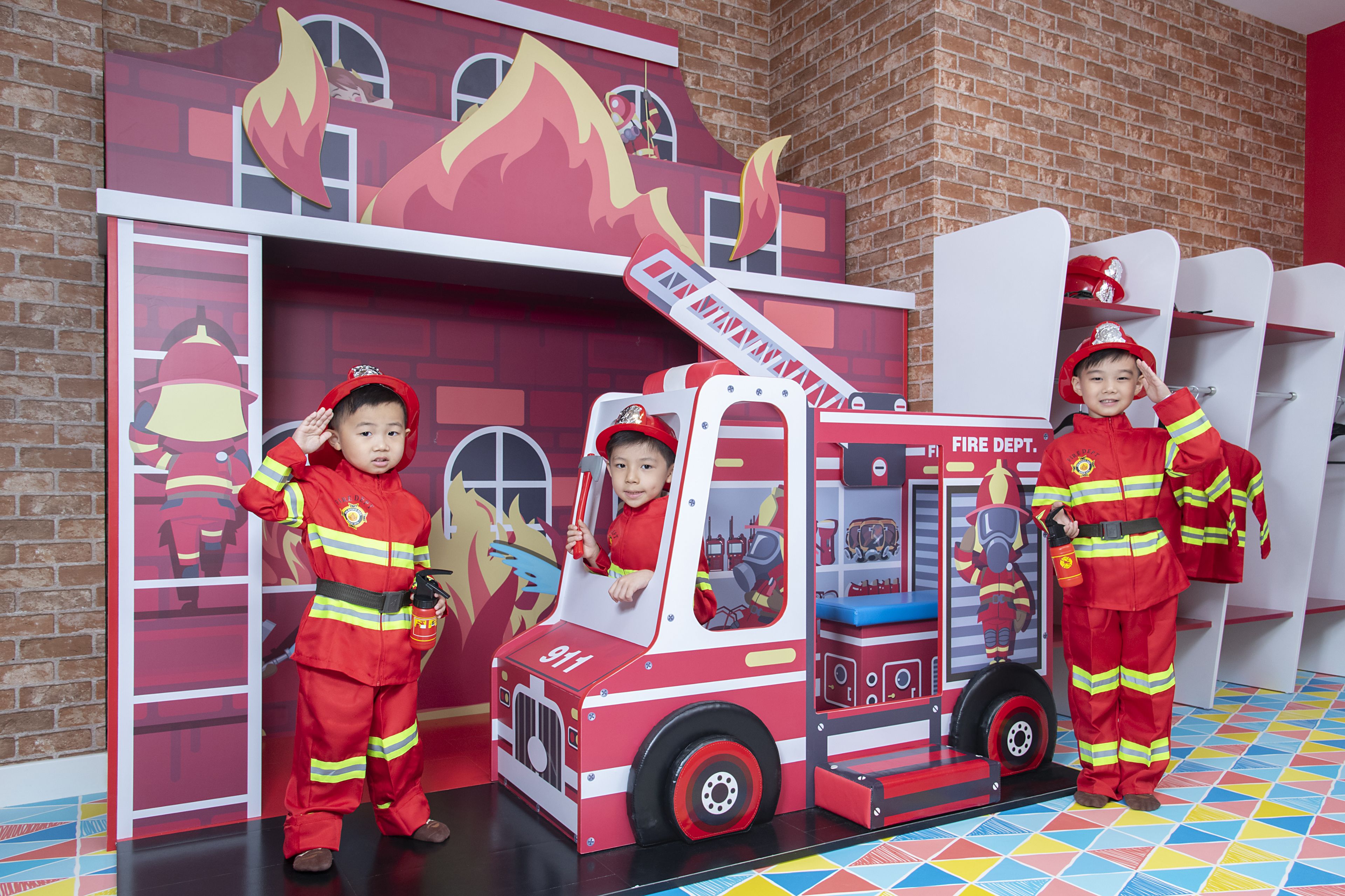 A play area with a fire truck playset, including a firefighter figure and a child in a firefighter costume.