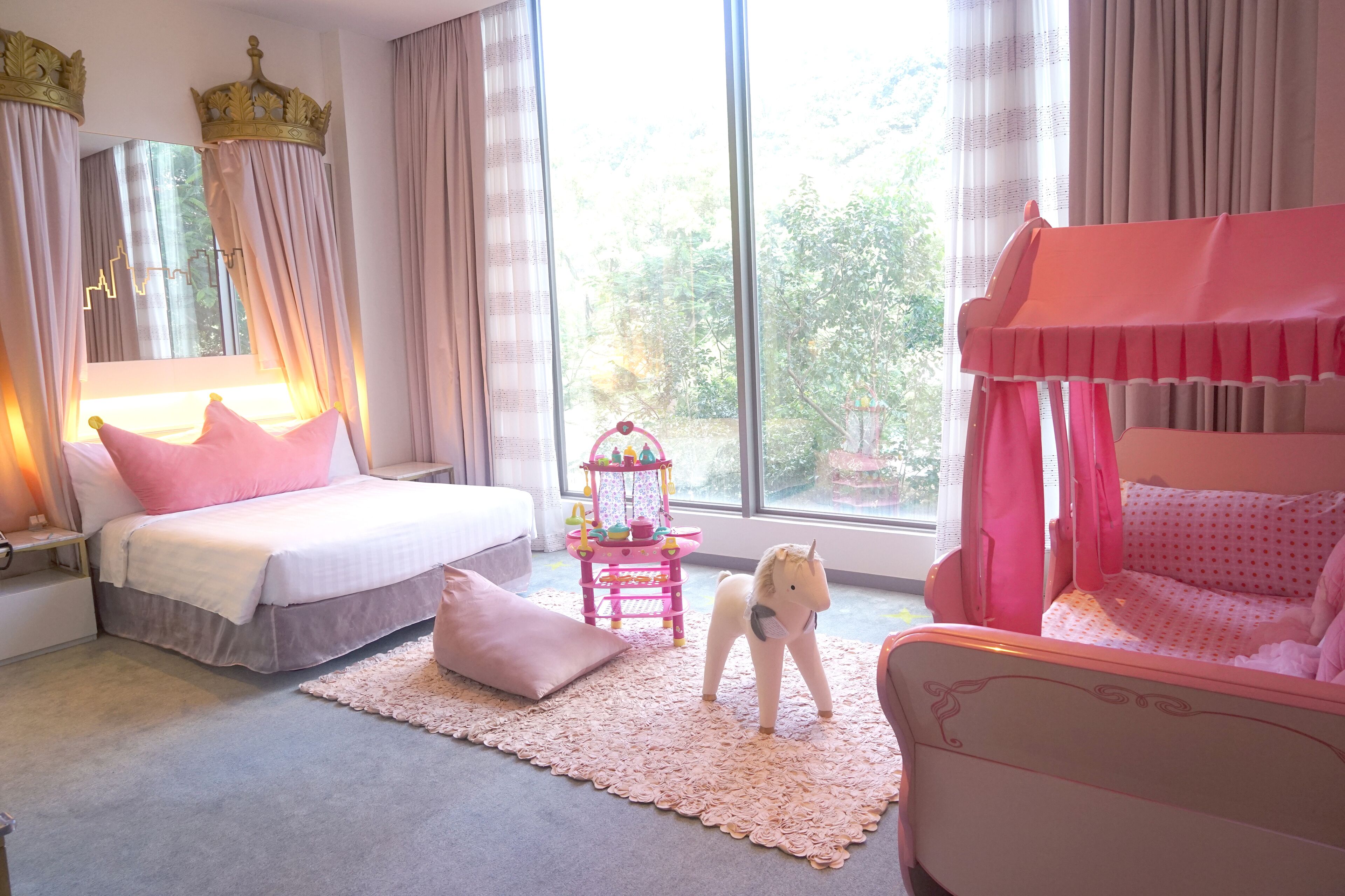 A bedroom with a large window, a bed, a crib, and a small toy horse.