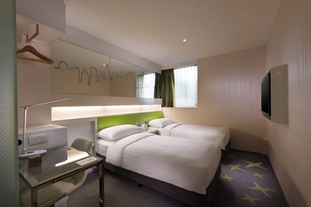 Et hotelværelse med to senge, et skrivebord med lampe, et spejl og et billede af en by skyline på væggen.