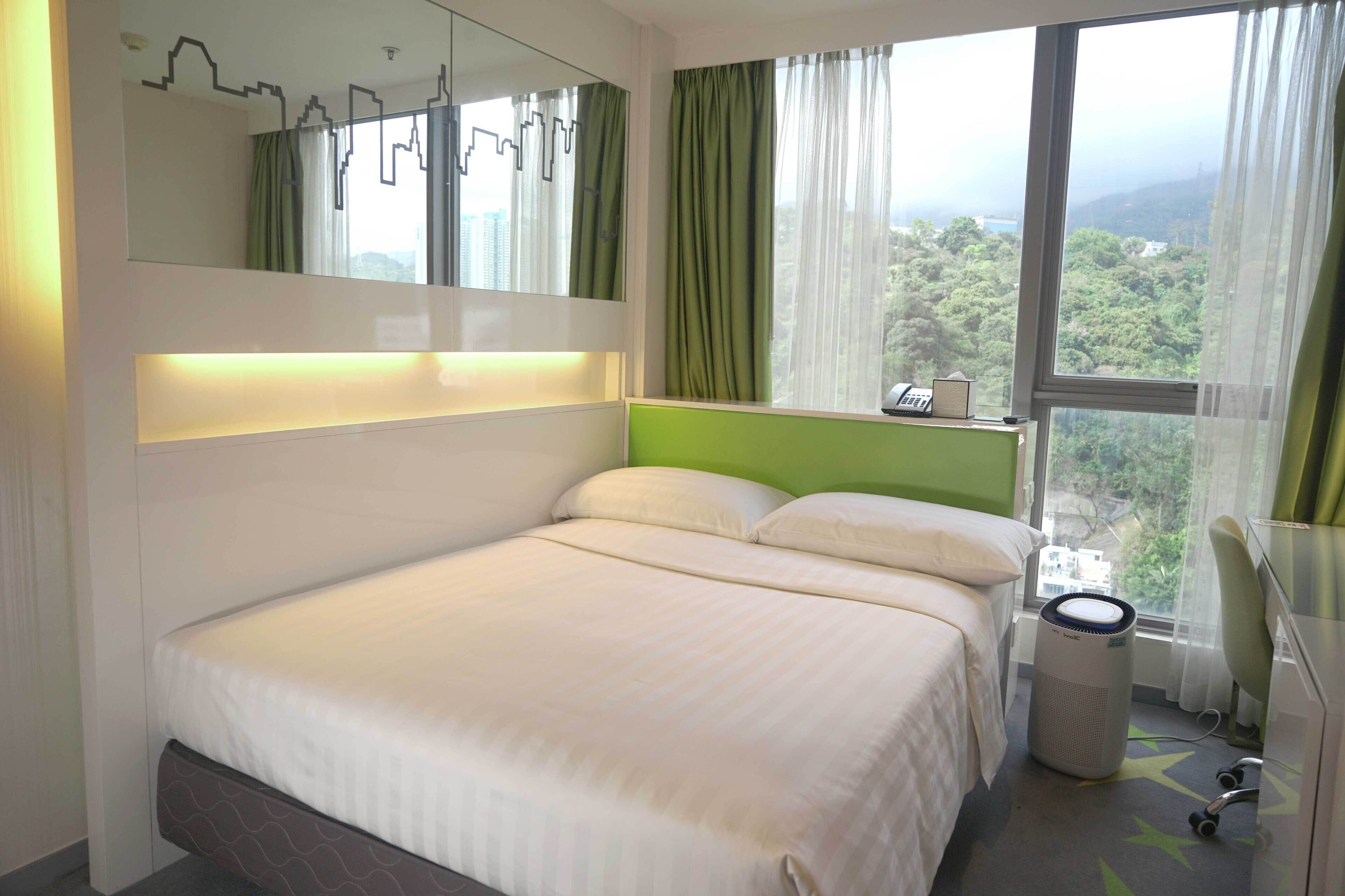 A hotel room with a bed, a bedside table, a mirror, a window with curtains, and a view of greenery.