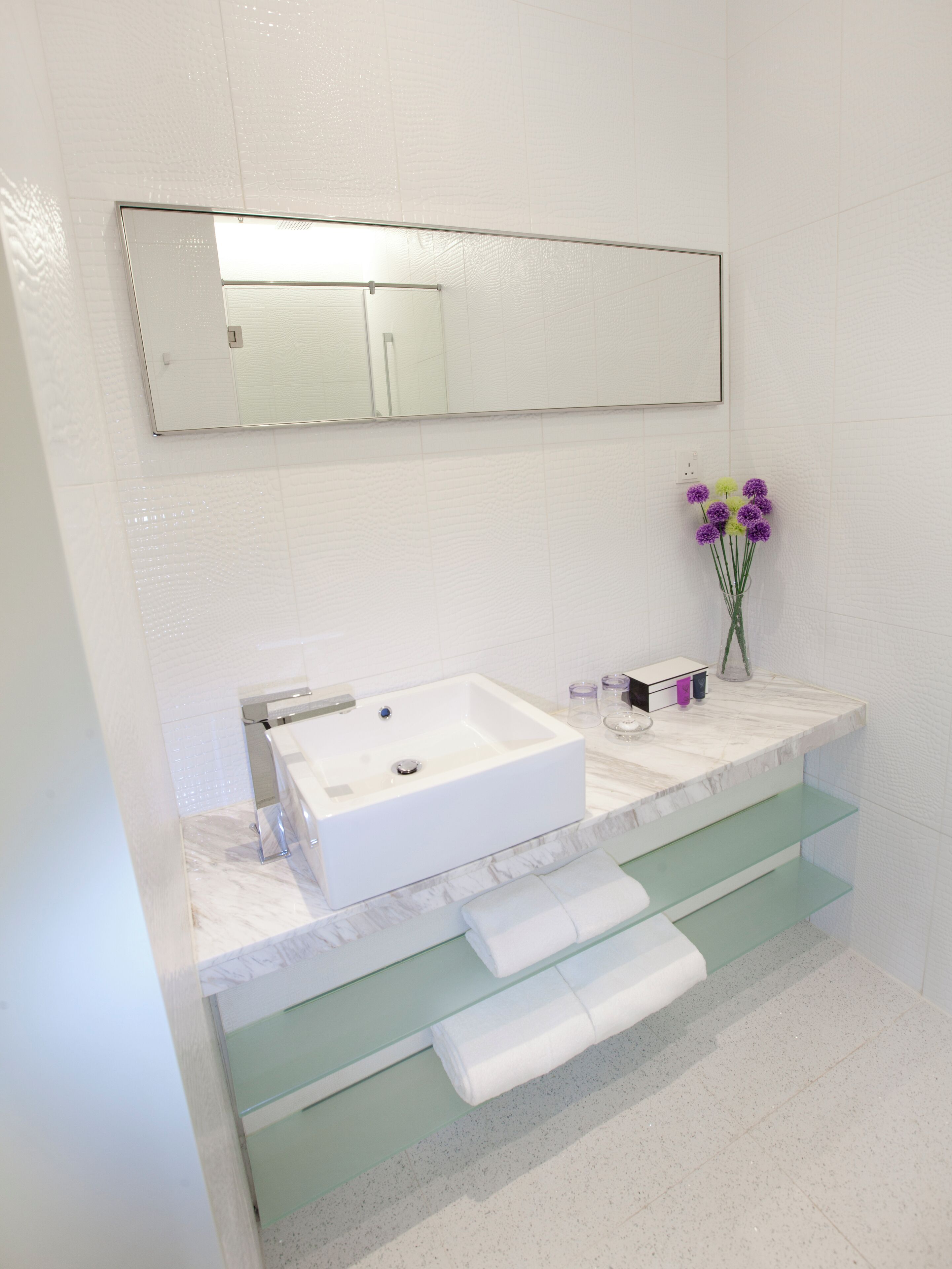 Un baño con un lavamanos blanco, un espejo encima y un florero con flores moradas en la encimera.