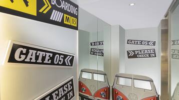 Un muro con indicazioni direzionali per 'GATE 05' e 'BOARDING M05', e il riflesso di un furgone d'epoca.