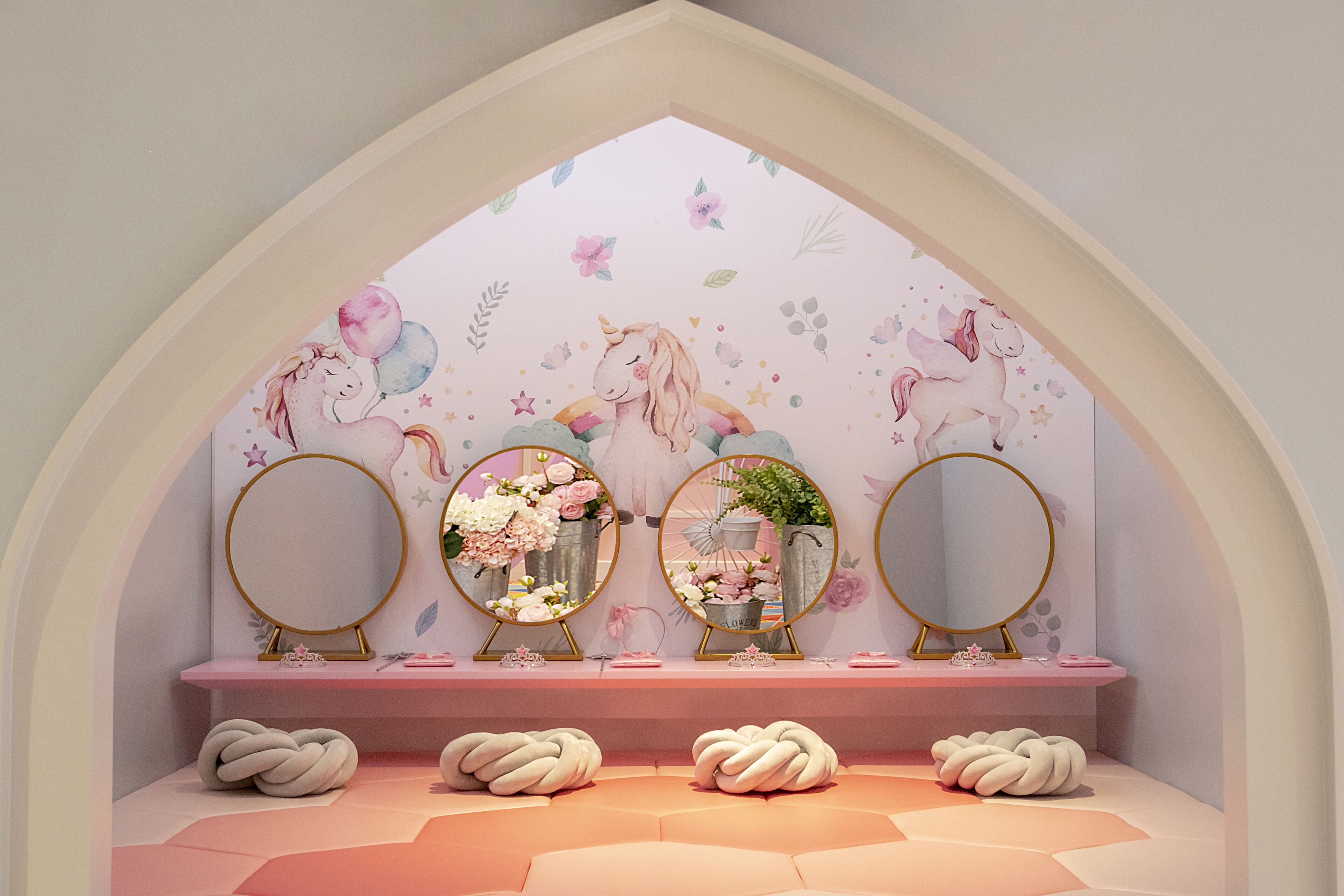 A room with a pink vanity, three round mirrors, and a floral mural on the wall.