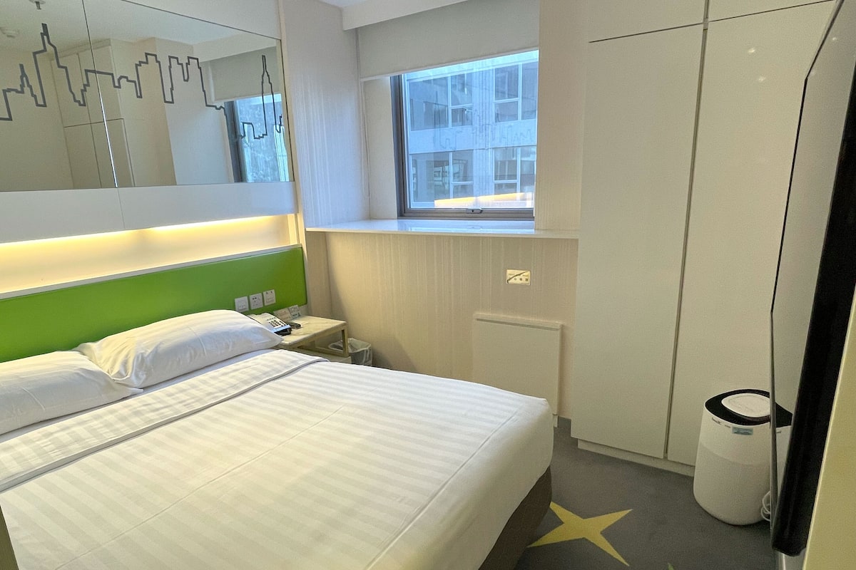 Et hotelværelse med en seng, et lille natbord, et spejl, et vindue og en radiator.
