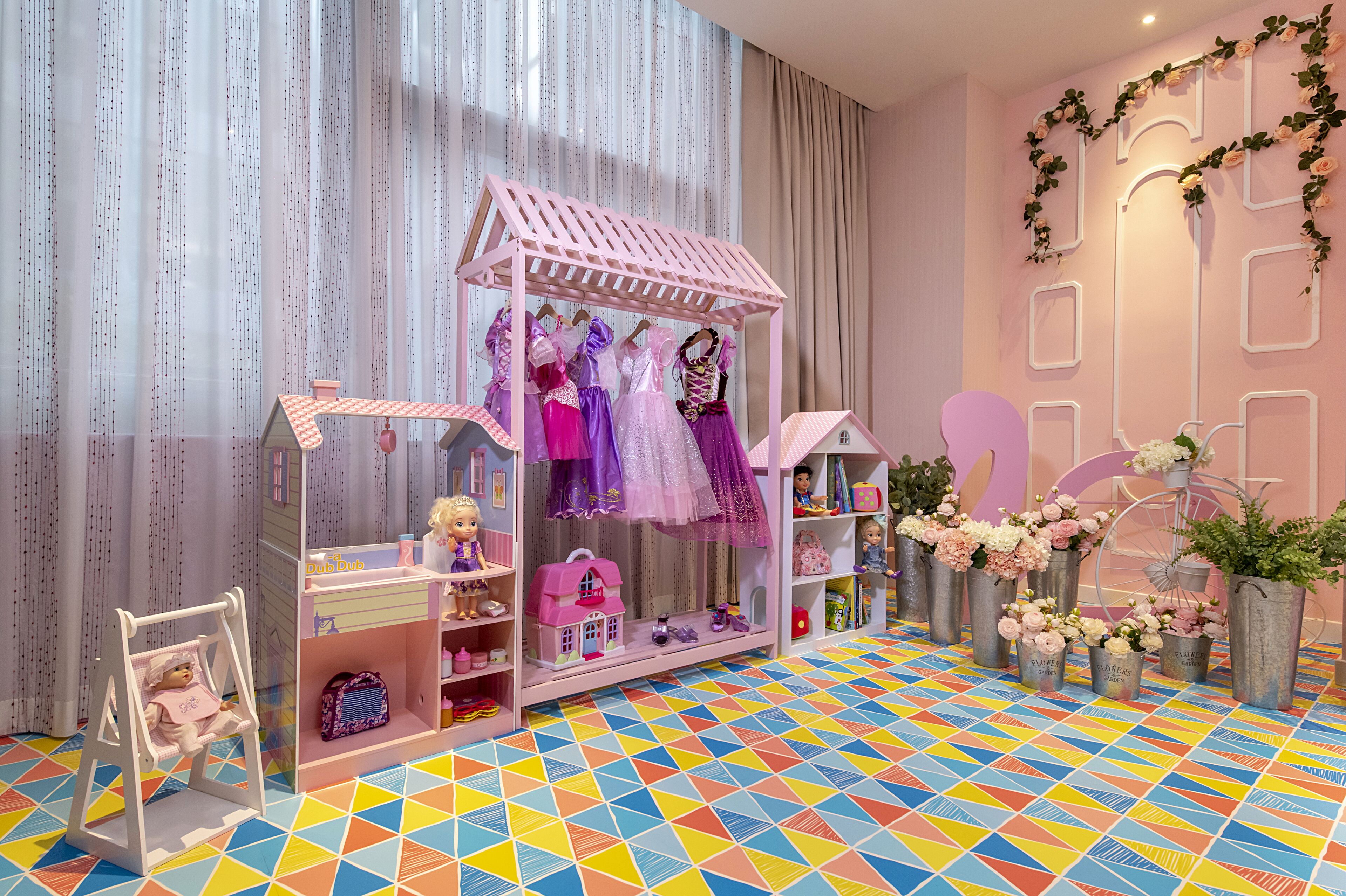 A dollhouse with a pink roof and various dolls inside, surrounded by vases of flowers and a colorful geometric floor pattern.