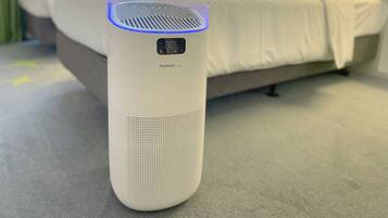 Een witte slimme speaker met een digitaal scherm op een tapijtvloer in een hotelkamer met een bed en groene muren.