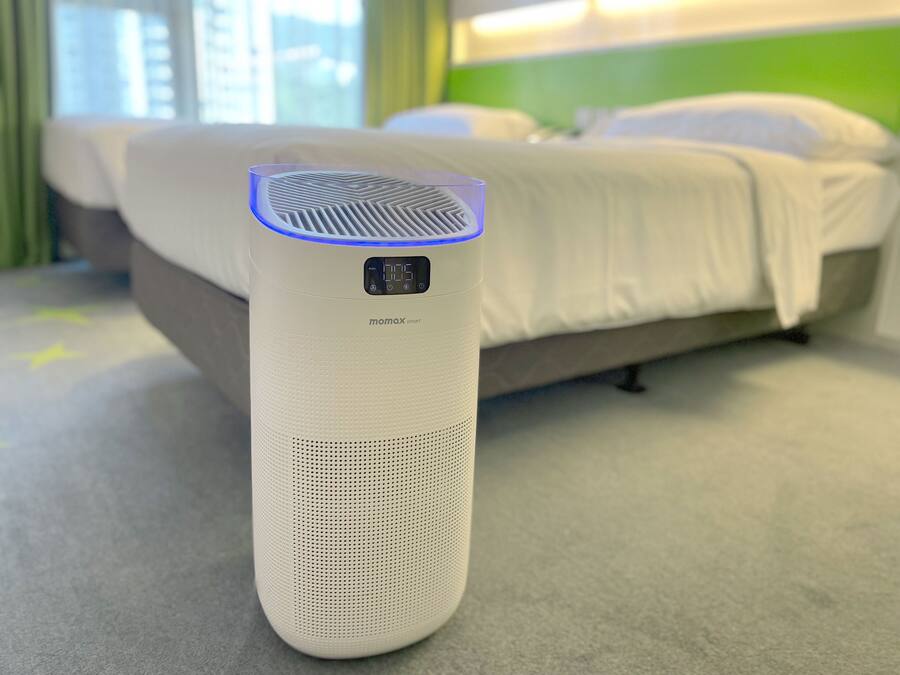 Een witte slimme speaker met een digitaal scherm op een tapijtvloer in een hotelkamer met een bed en groene muren.