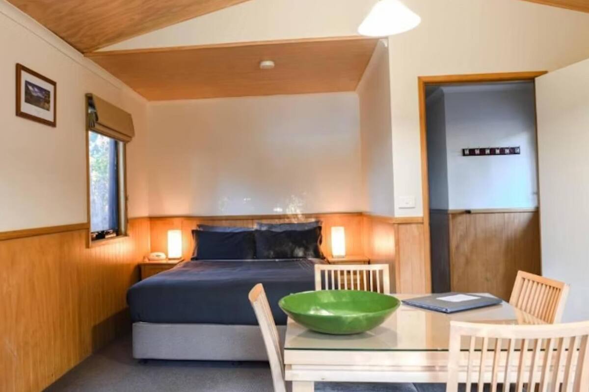 A compact bedroom with a bed, bedside lamps, a small table with a green bowl, and a door.