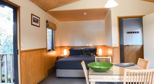 A compact bedroom with a bed, bedside lamps, a small table with a green bowl, and a wooden door.