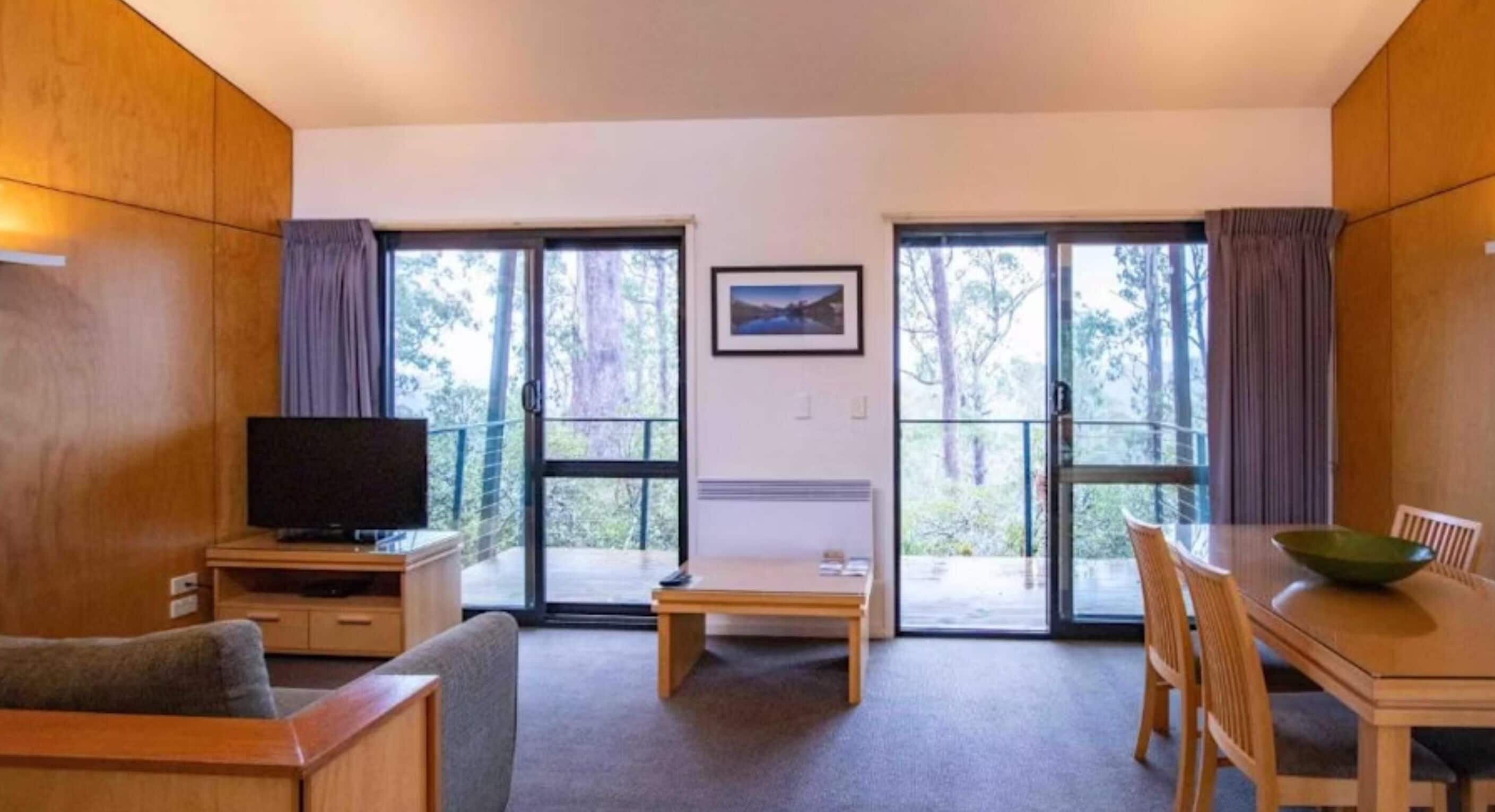 A room with a TV, a dining table with chairs, and a bench. There are two sliding glass doors leading to a balcony.