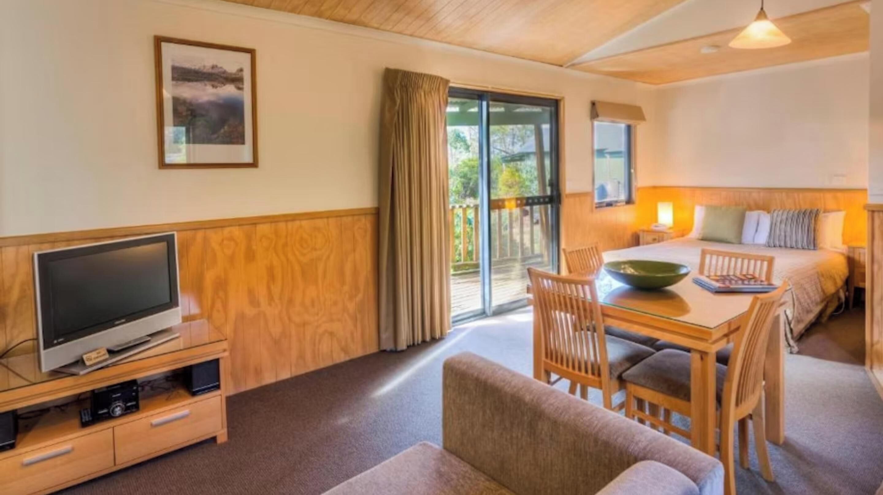 A cozy room with a bed, a dining table with chairs, a TV on a wooden stand, and a large window with curtains.
