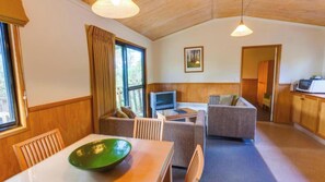 A cozy room with a wooden ceiling, a dining table with a green bowl, a sofa, a television, and a kitchen area with a microwave.