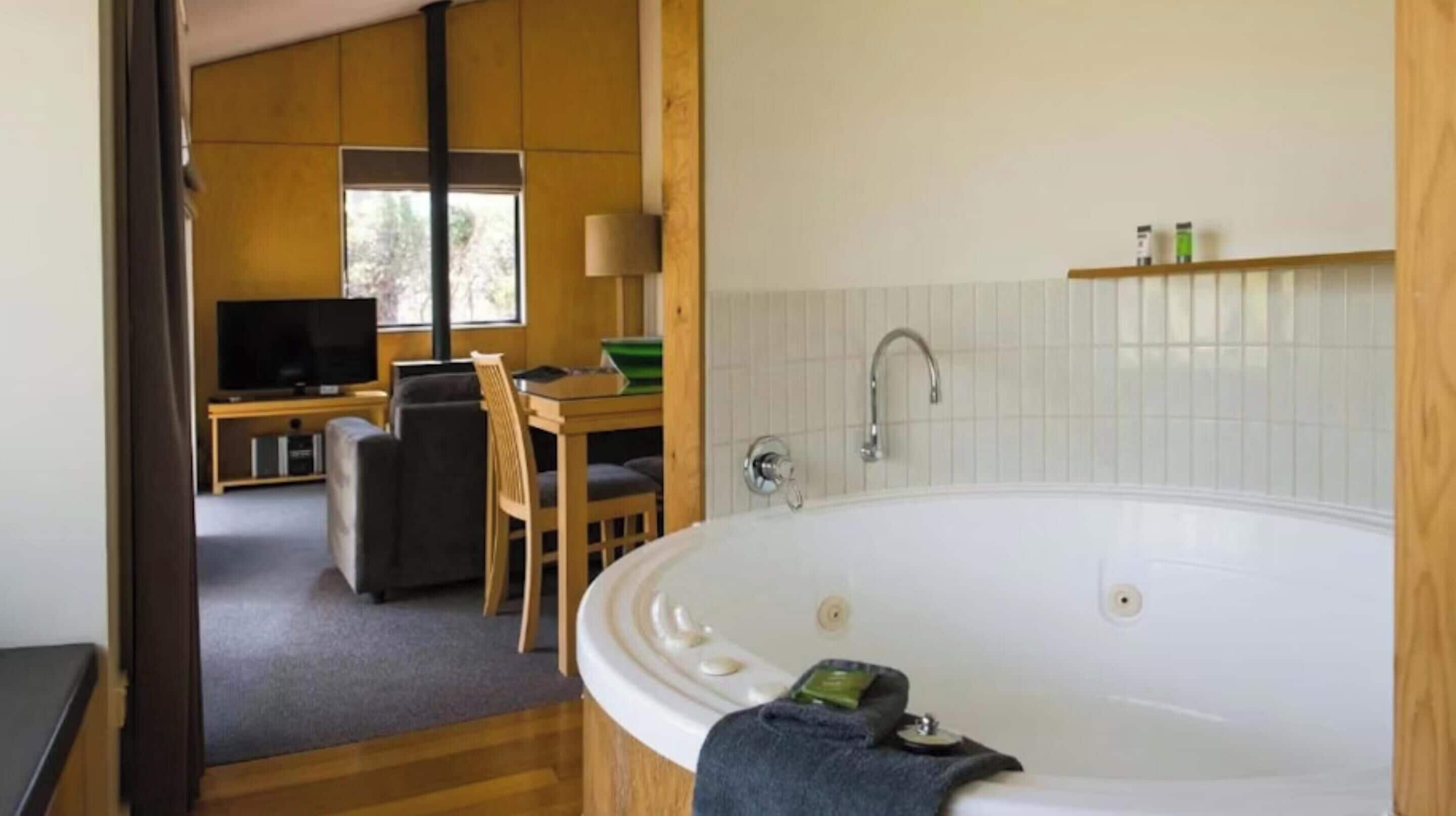A modern bathroom with a large bathtub, a wooden chair, a small table, and a television in the background.