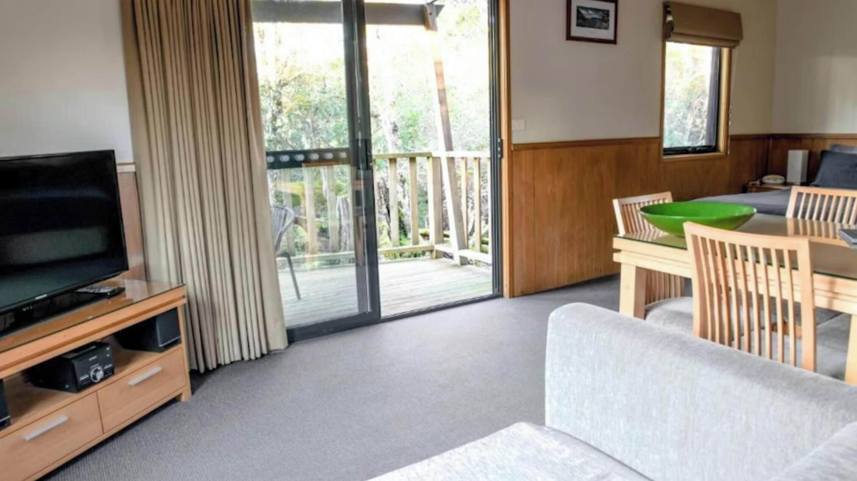 Una habitación con televisión sobre un soporte de madera, un sofá, una mesa de comedor con sillas y un ventanal corredero que da a una terraza exterior.