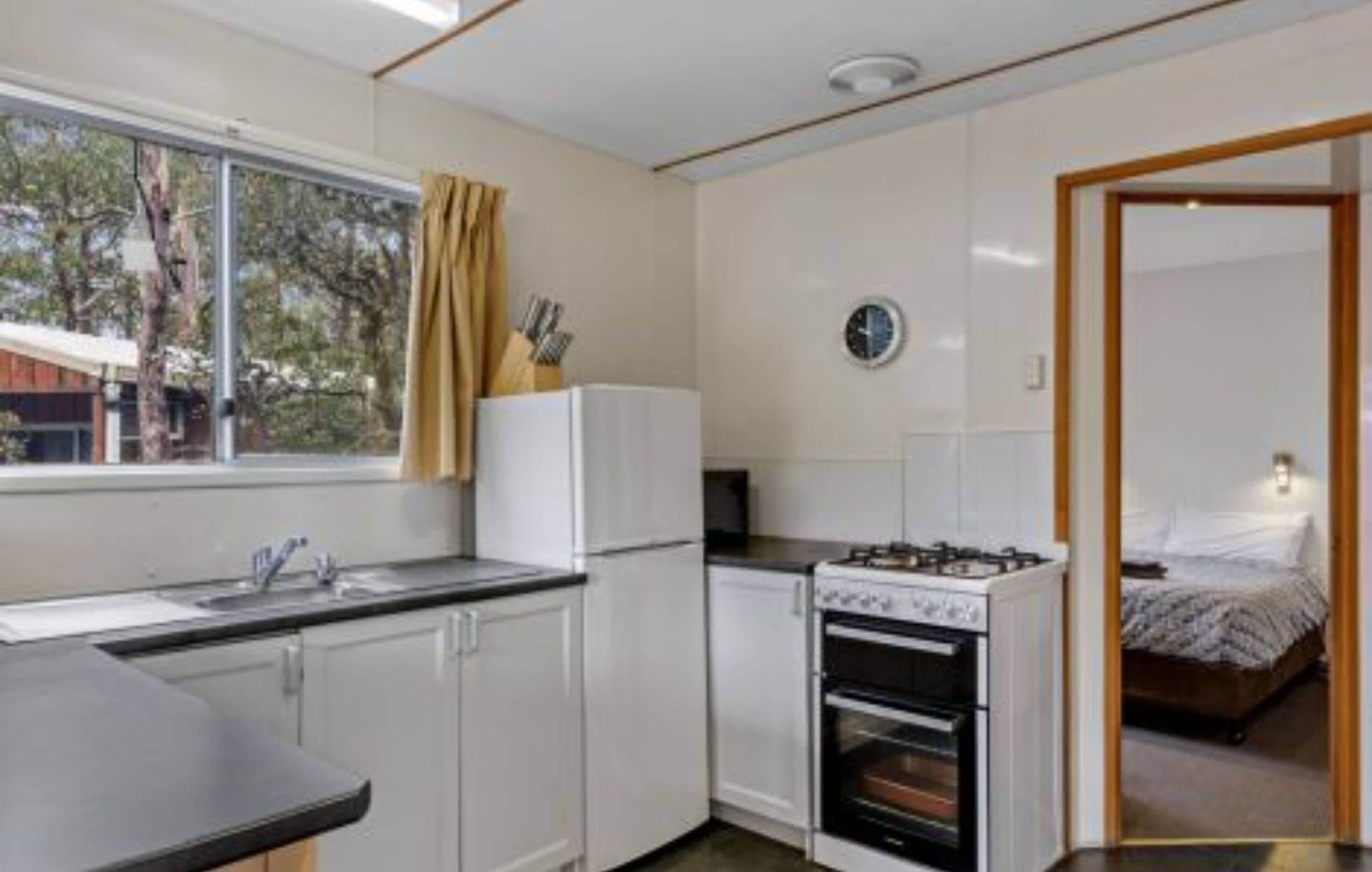 A compact kitchen with a stove, oven, and sink. A bed is visible through an adjacent door.