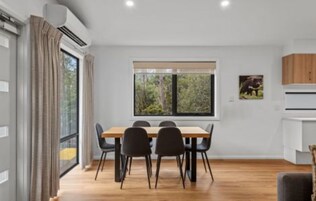 Un comedor moderno con una mesa y sillas de madera, un ventanal con vistas a zonas verdes, y una cocina blanca con armarios.