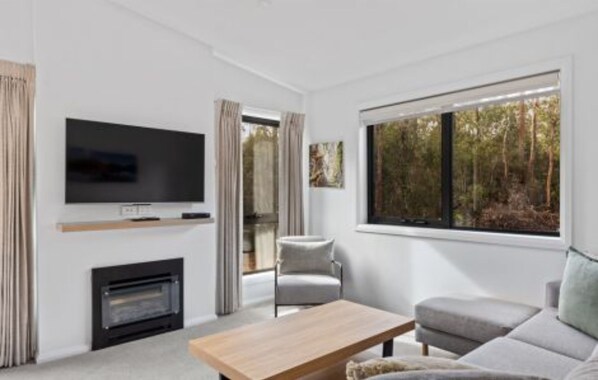 Una sala de estar moderna con televisor de pantalla plana, chimenea, sofá y ventanas amplias con vistas a un bosque.