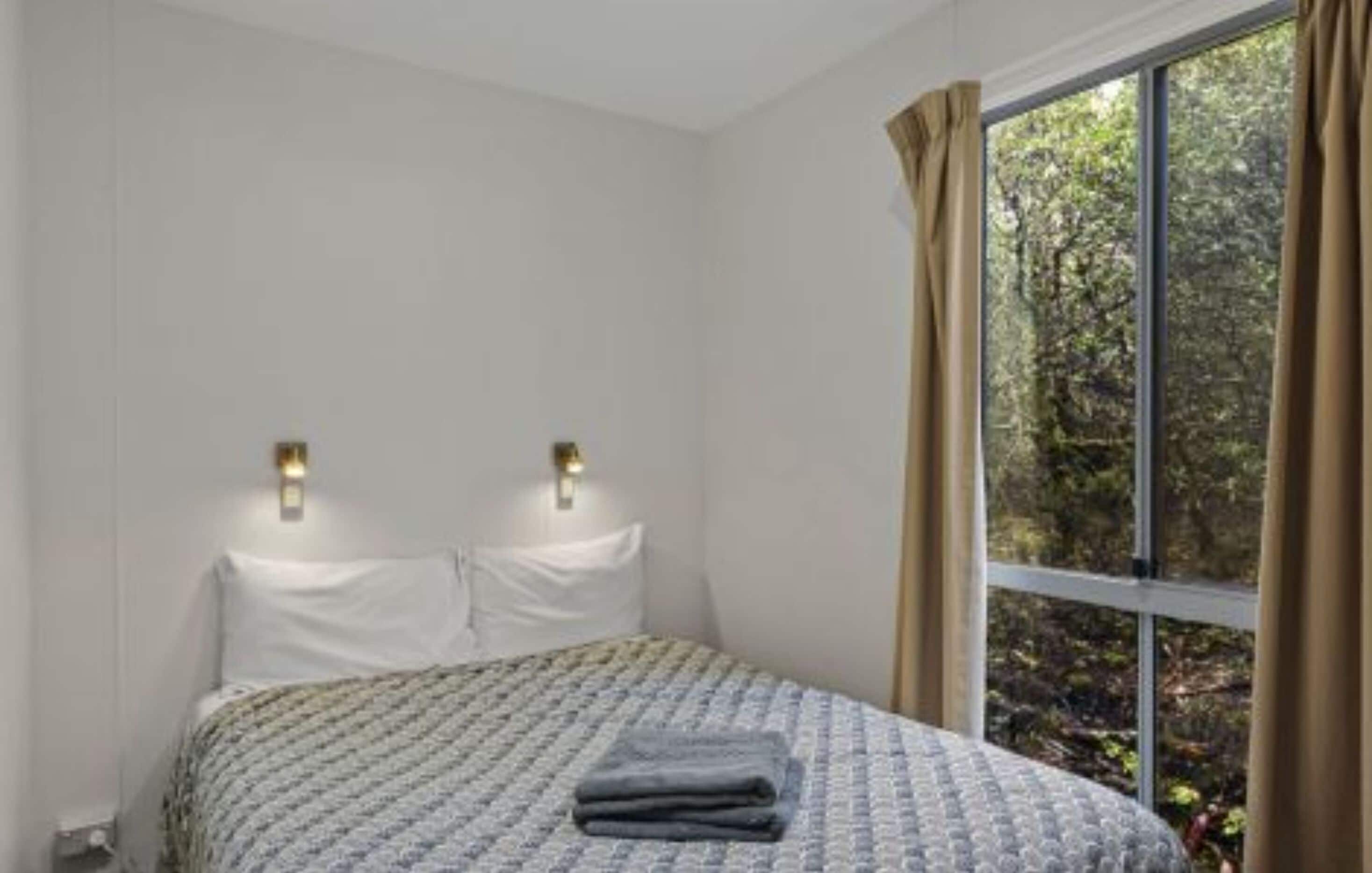 A bedroom with a bed, two wall-mounted lights, a window with curtains, and a view of trees.