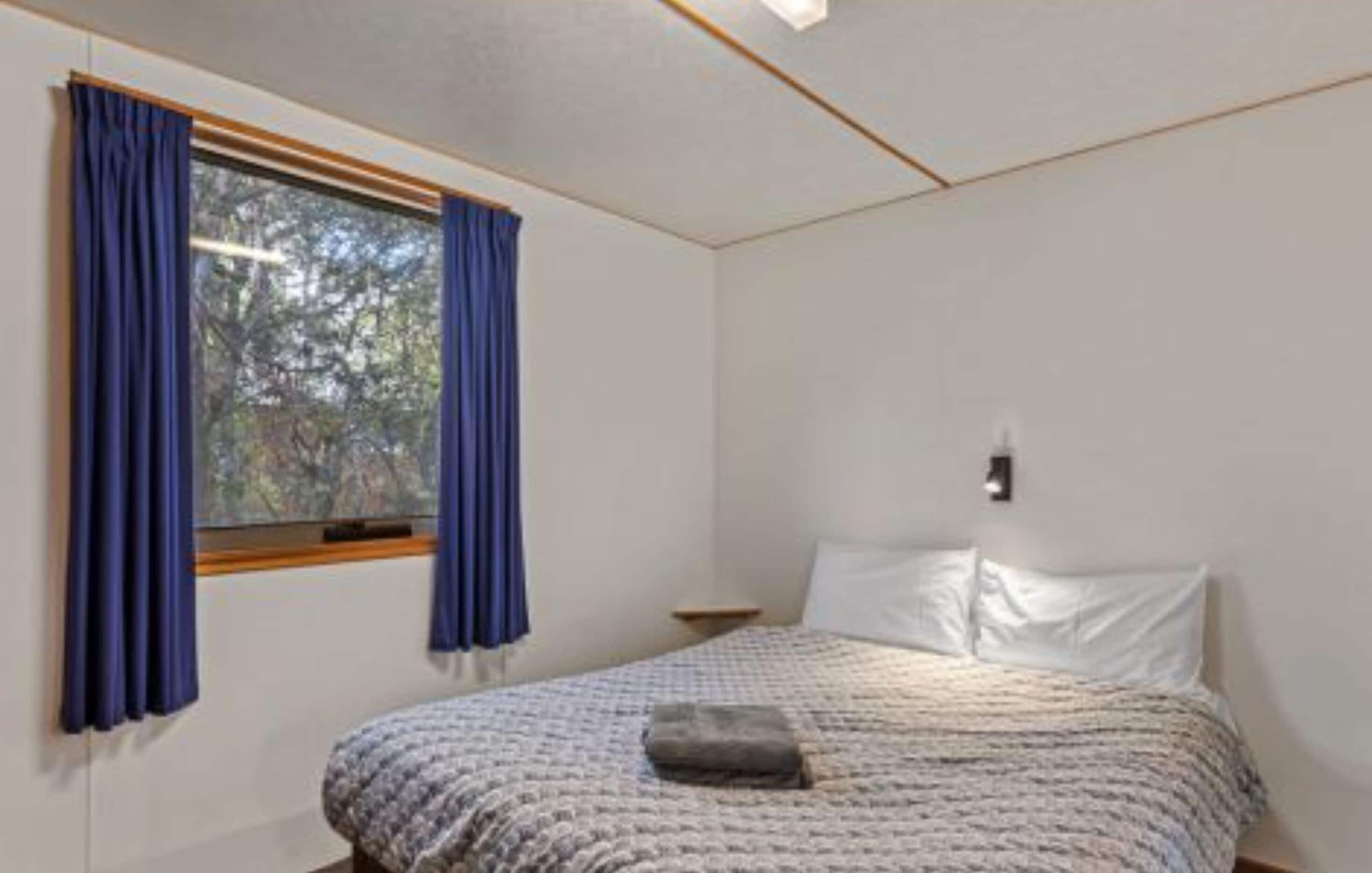 A bedroom with a bed, a window with curtains, a bedside table, and a wall-mounted light.