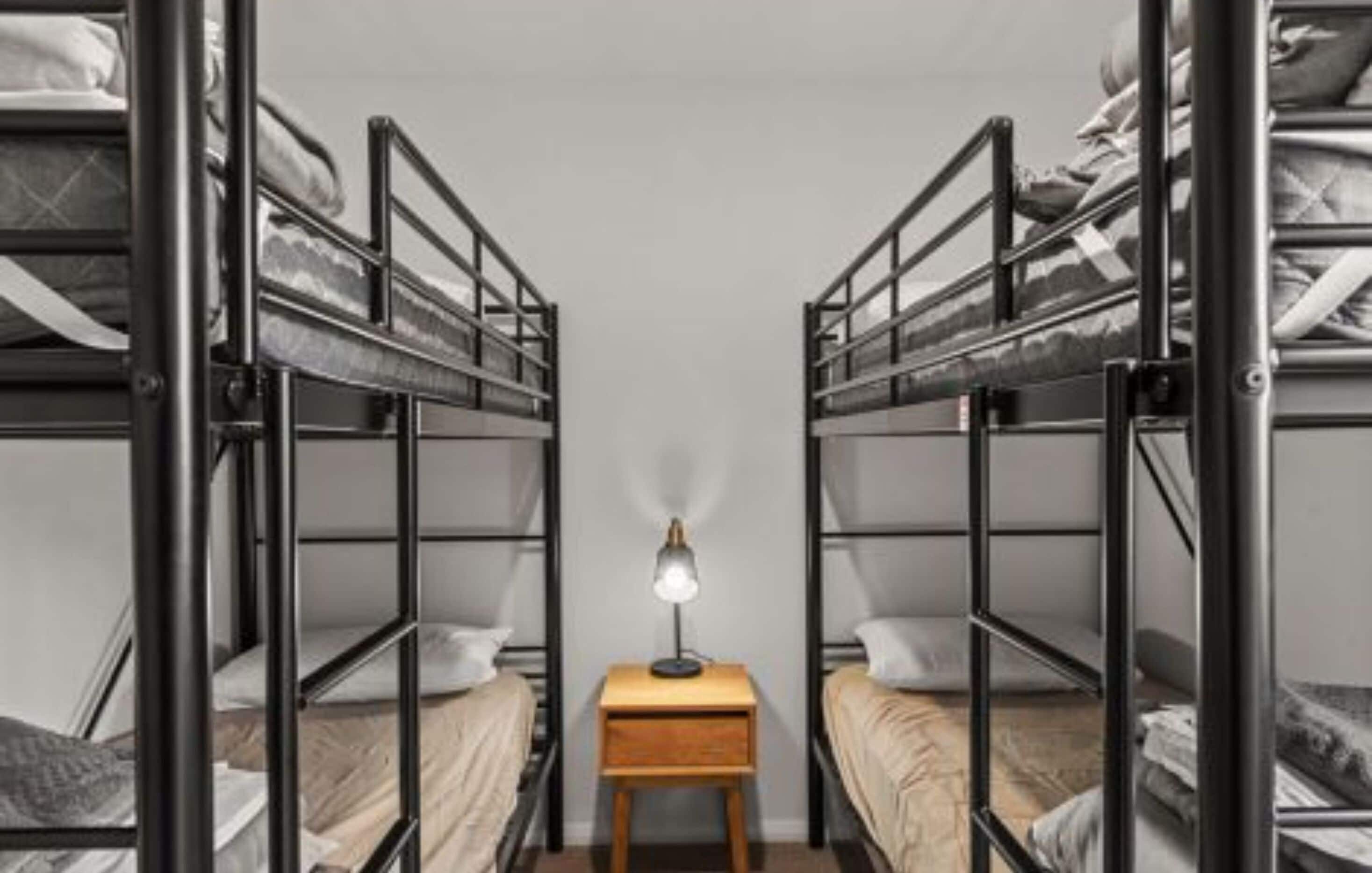 Habitación con literas de metal a ambos lados, una cama individual en el centro y una mesita de noche con lámpara.