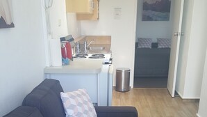 A modern living room with a dark sofa and patterned cushions, a small kitchen with a stove and sink, and a bedroom visible through an open door. - Bayview Motel (Paihia)