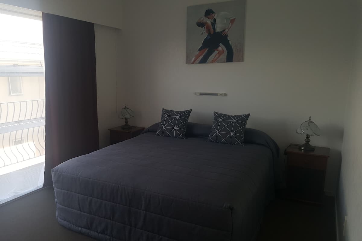A bedroom with a bed, two bedside tables with lamps, a painting on the wall, and a window with a view of buildings.