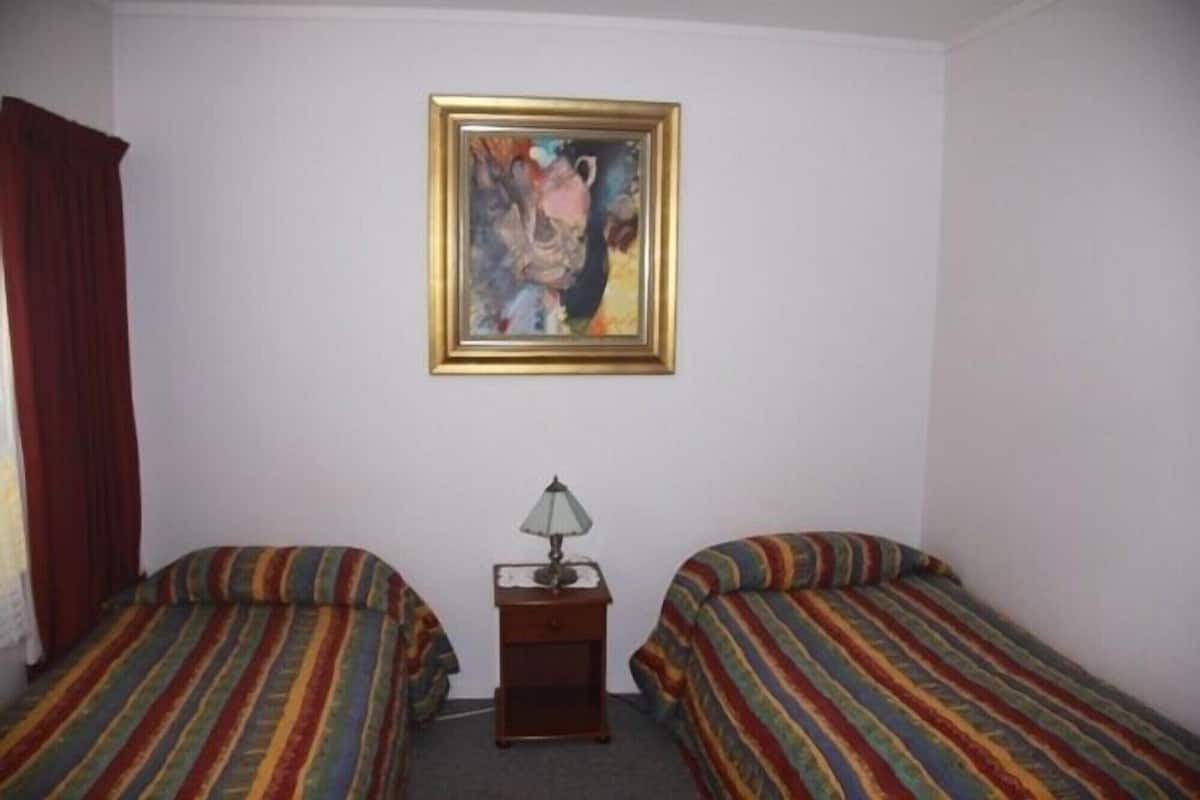 A room with two single beds, a small wooden nightstand with a lamp, and a framed painting on the wall.