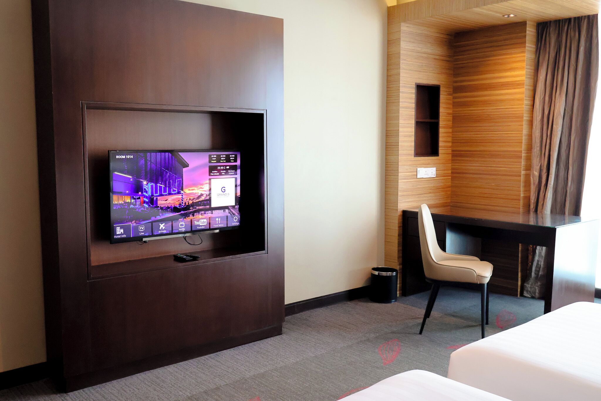 A hotel room with a flat-screen TV mounted on a dark wooden wall, a desk with a chair, and a bed.
