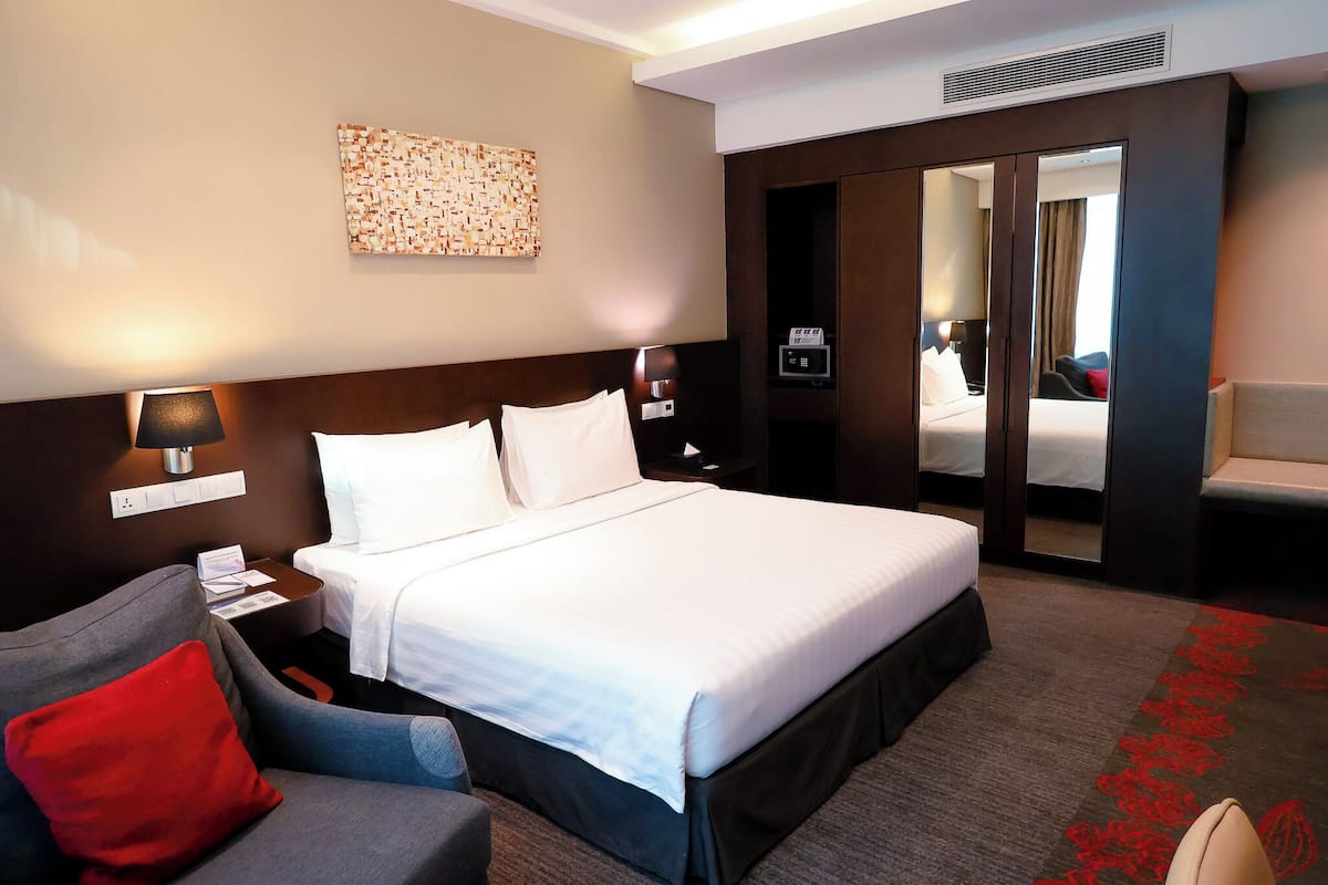 A hotel room with a large bed, a desk with a lamp, a sofa with a red pillow, a wall-mounted artwork, and a mirror.
