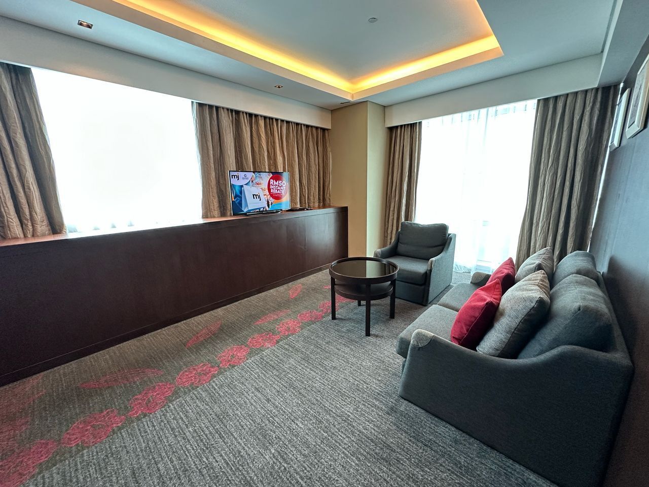 Una habitación de hotel con sofá, sillón, una mesita y televisor montado en la pared.
