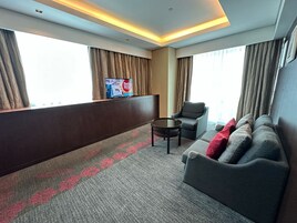 Una habitación de hotel con sofá, sillón, una mesita y televisor montado en la pared.