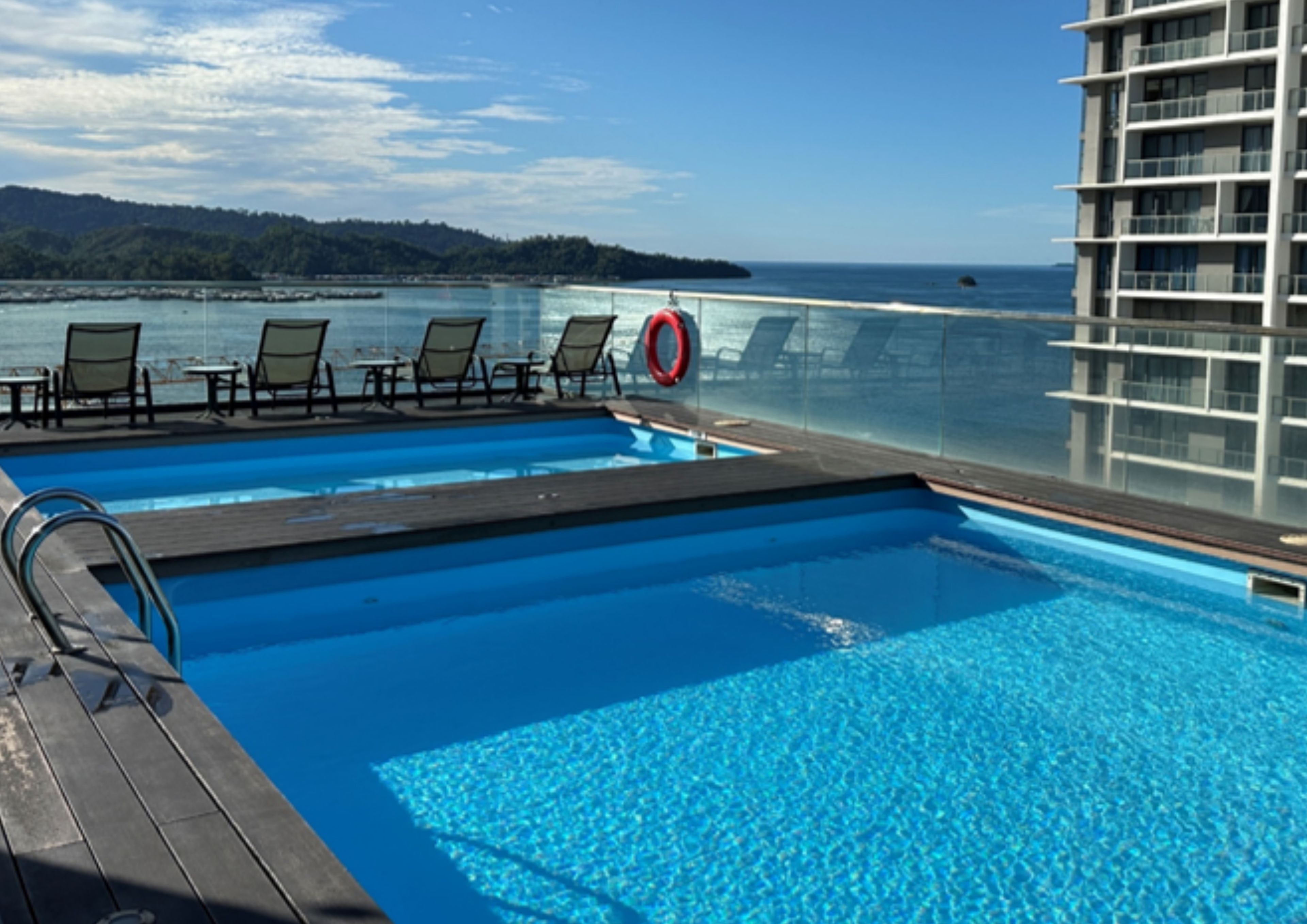 A pool area with lounge chairs and a lifebuoy, overlooking a body of water and a distant landmass.