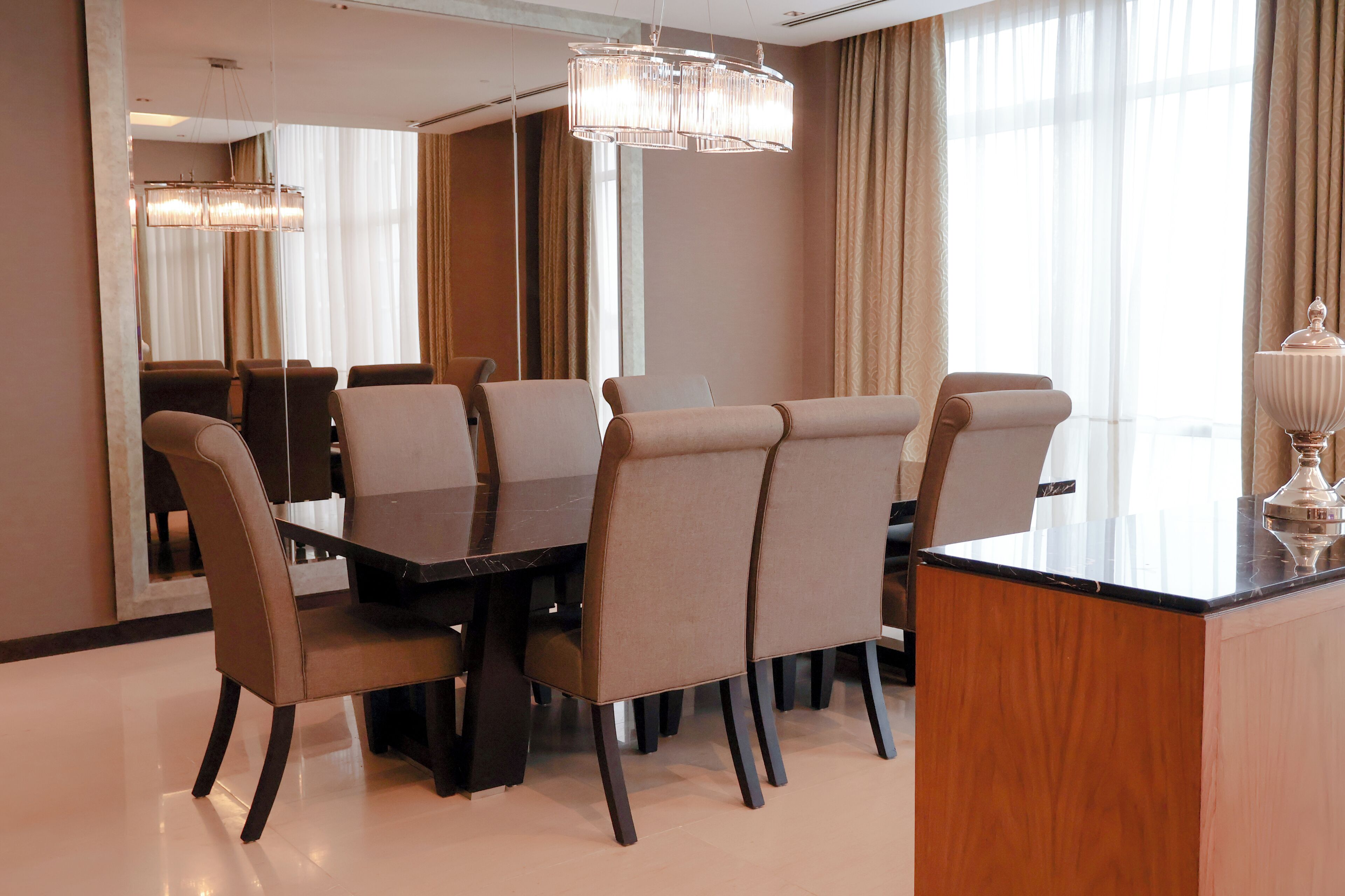 A modern hotel dining room with a rectangular table, six chairs, a large mirror, and a chandelier.