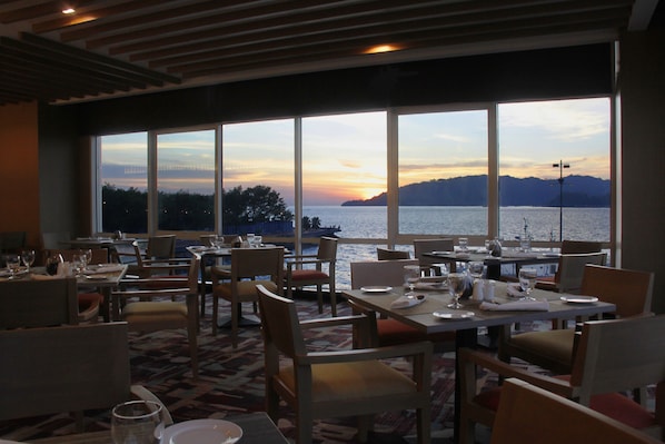 A restaurant with tables and chairs arranged for dining, large windows offering a view of the water and sunset.