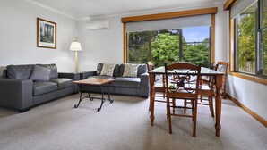 A living room with a grey sofa, a wooden dining table, and chairs, a lamp, and a framed picture on the wall. - Marsden Court Apartments (Strahan)
