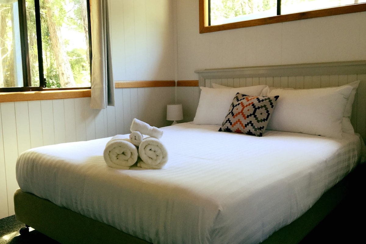A neatly made bed with white linens, a patterned pillow, and rolled towels on the side.