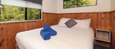 Un dormitorio con una cama grande, paneles de madera, dos ventanas con vistas a la vegetación y una mesita de noche pequeña.