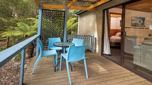Terraza de madera con sillas azules y una mesa, pared de piedra y puerta de cristal que da a una habitación con cama y un cuadro.