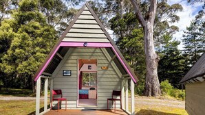 Una pequeña estructura de madera triangular con un porche y dos sillas, rodeada de árboles.