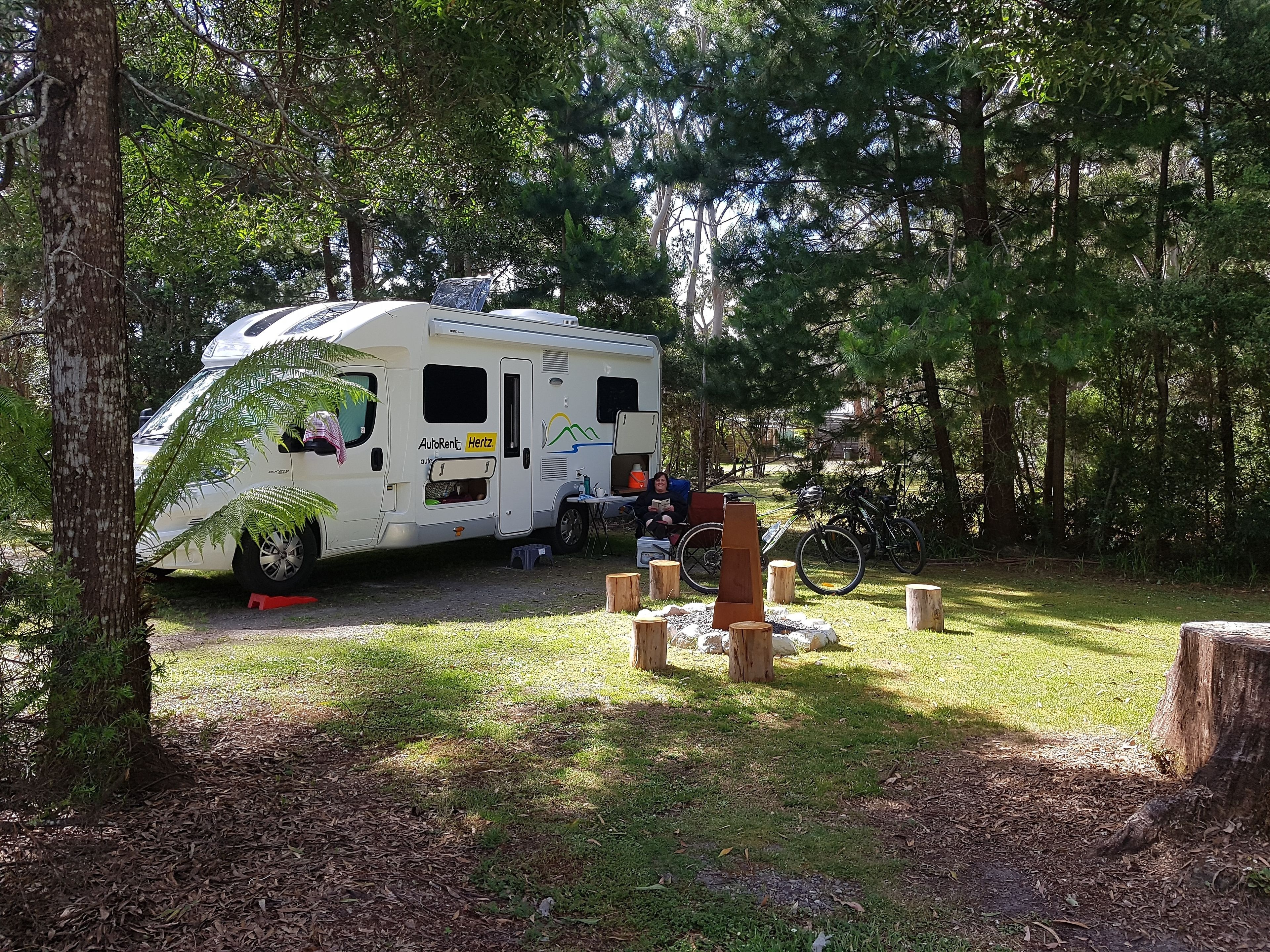 A white RV parked in a wooded area with a picnic table and a bicycle nearby.