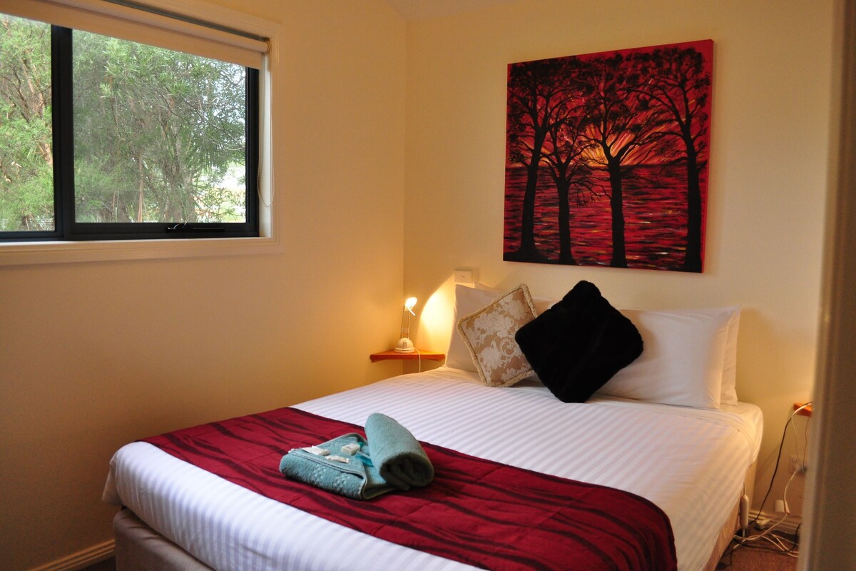 A hotel room with a bed, a window, a painting, and a bedside lamp.
