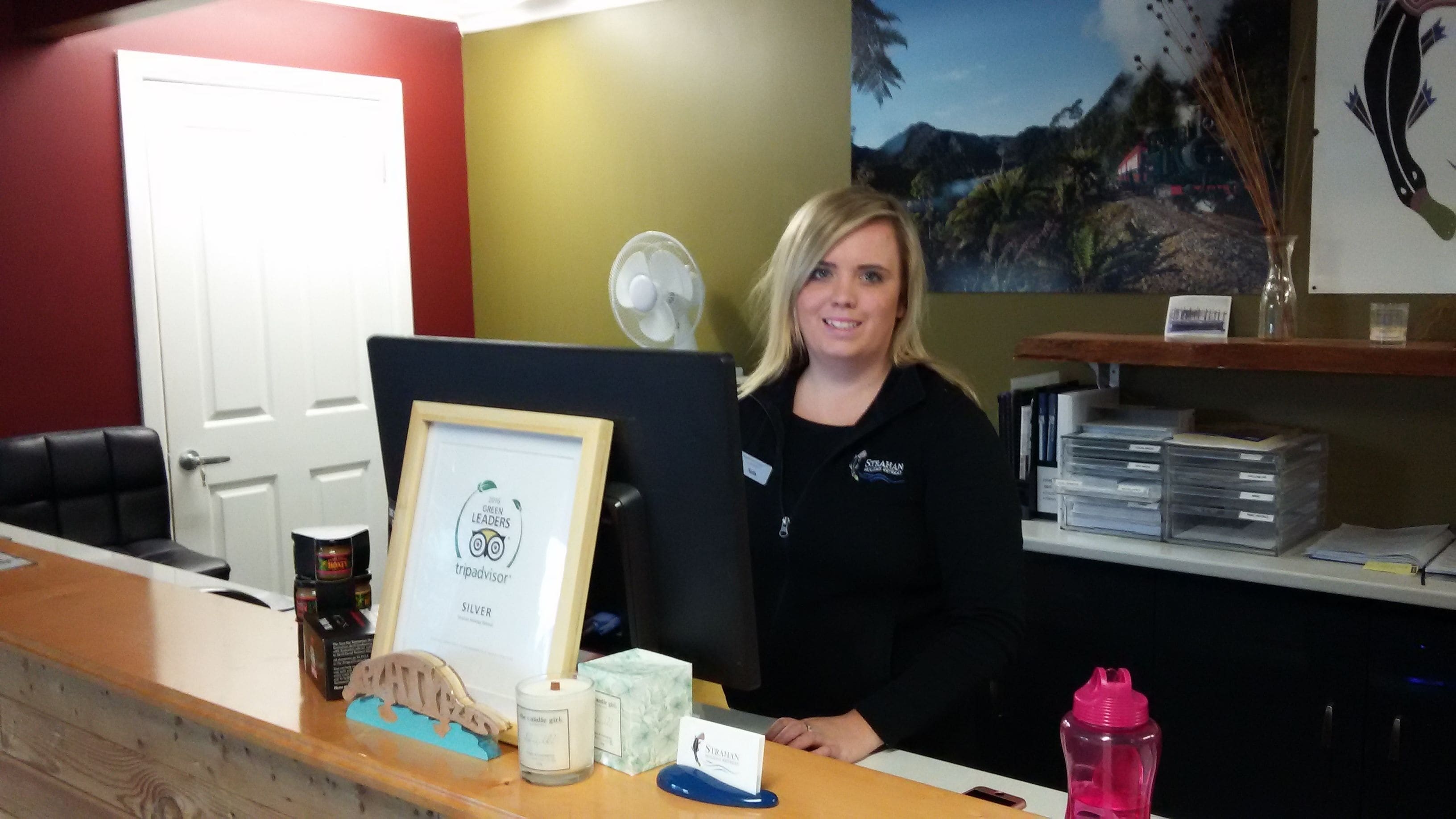 A reception desk with a framed certificate, a small candle, a water bottle, and a fan in the background.