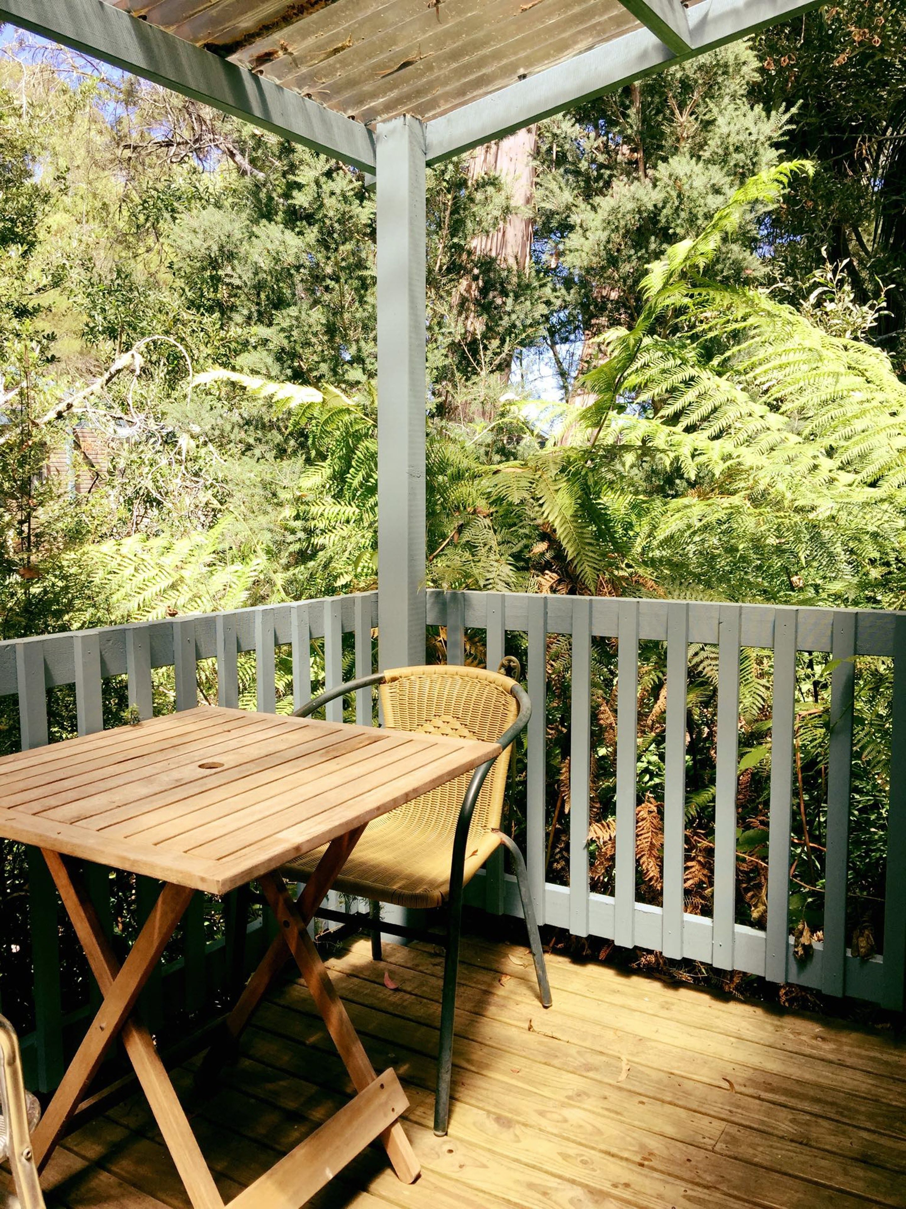 Een houten terras met een tafel en stoelen, omgeven door een witte balustrade, met uitzicht op een weelderige tuin met verschillende planten en bomen.